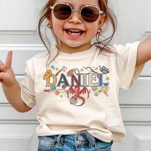 May include: A cream-colored t-shirt with the name "DANIEL" in a colorful, fantasy-themed design. The design includes a red dragon, dice, stars, and various fantasy elements. The shirt is worn by a child.