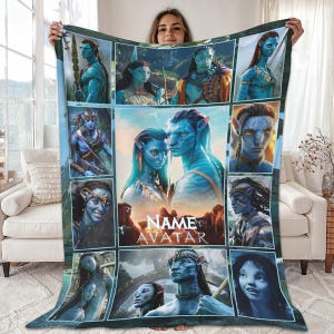 Puede incluir: Una manta de forro polar azul con múltiples imágenes de personajes de la película Avatar. La manta tiene una imagen central de dos personajes Na'vi con el texto "NAME AVATAR" debajo. La manta está sostenida por una persona.