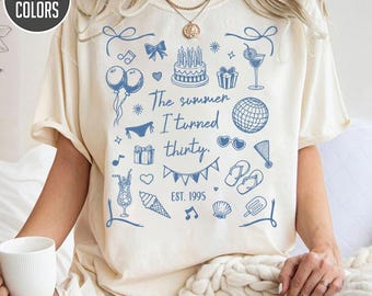 May include: Cream-colored t-shirt with a blue summer-themed graphic. The design includes balloons, a cake, a bikini, and the text "The summer I turned thirty. EST. 1995." The shirt is from Comfort Colors.
