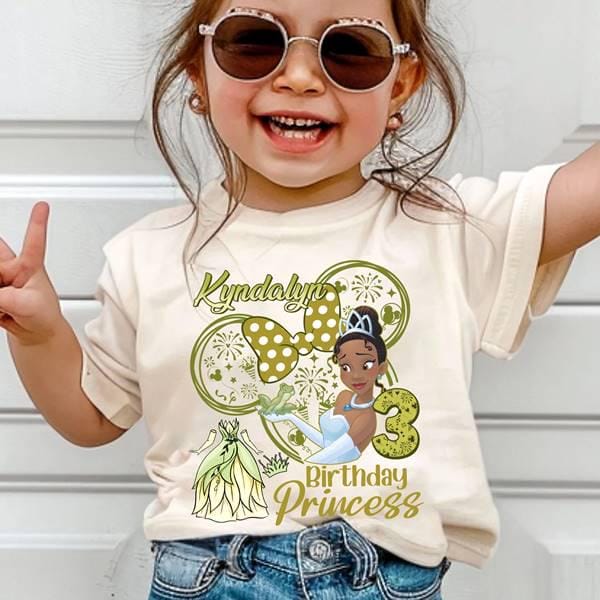 May include: A cream-colored t-shirt featuring a graphic of a princess with a crown, a number 3, and the words "Birthday Princess." The design includes a bow and the name "Kyndalyne" in a script font. The shirt is worn by a young child.