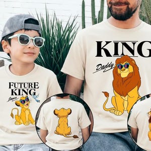 May include: Beige t-shirts with lion graphics. One shirt says "KING Daddy" with a lion wearing sunglasses. The other says "FUTURE KING" with a lion and a bird. The back of the shirts also have lion graphics.