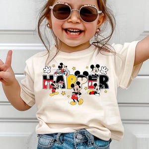 A young girl wearing sunglasses and a Mickey Mouse-themed t-shirt is smiling and making a peace sign gesture.