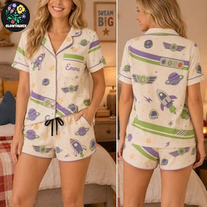 May include: A cream-colored pajama set featuring a short-sleeved button-down shirt and matching shorts. The fabric is decorated with a space-themed pattern of rockets, planets, and stars in purple and green. The shirt pocket displays the name Emma.