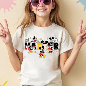 A young girl wearing a white t-shirt with the word "HAPPY" and various Disney characters, including Mickey Mouse, is smiling and making a peace sign gesture.