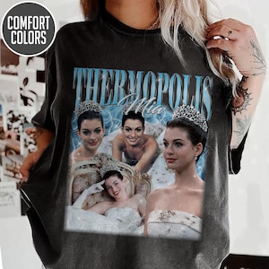 May include: Dark gray t-shirt featuring a collage of images with the text "Thermopolis Mia" in light blue. The images depict a woman wearing a tiara and formal attire. The shirt has a vintage, distressed look.