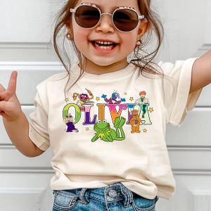 May include: A cream-colored t-shirt featuring the name "OLIVIA" in a colorful, playful font, with Muppet characters and stars. The design includes Kermit the Frog and other Muppet characters. The shirt is worn by a child.