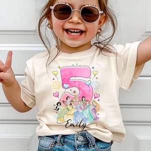 May include: Cream-colored t-shirt featuring a large pink number 5 with a watercolor effect, surrounded by hearts and stars. Below the number, a group of cartoon princesses is depicted. The name "Emily" is written in cursive below the princesses.
