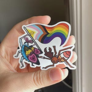 Puede incluir: Una pegatina con una polilla y una hormiga de dibujos animados sosteniendo una bandera del Orgullo. La polilla tiene alas azules, amarillas y rosas con diseños de corazón. La hormiga es marrón con rayas rojas y naranjas. La bandera es de arcoíris.