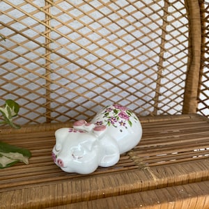 May include: A white ceramic pig ornament with pink details, such as a bow, ears, and snout. The pig is decorated with purple floral patterns and is lying down. The ornament is placed on a woven surface.