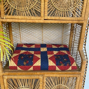 May include: A handmade quilt with a patchwork design in red, navy blue, and tan. The quilt is displayed on a vintage wicker shelf unit with a woven pattern. The quilt features floral embroidery.