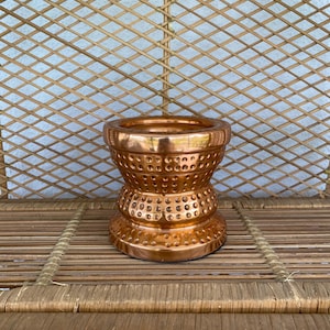 May include: A copper-coloured decorative candle holder with a unique hourglass shape. The candle holder has a wide, open top and a base with a series of small, evenly spaced holes. The surface has a shiny, metallic finish.