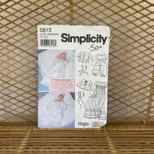 May include: A vintage Simplicity sewing pattern, size A, for baby clothes. The pattern includes illustrations of a dress, jacket, and bonnet. The pattern is set against a woven wicker background.