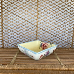 May include: Square ceramic dish with a pale yellow interior and a light blue rim. The dish is decorated with pink and purple roses and blue floral accents. The dish is sitting on a woven surface.