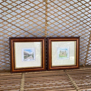 May include: Two framed watercolour prints with brown wooden frames. The prints depict architectural scenes with detailed brushstrokes. The frames are displayed against a woven wicker background, adding a touch of rustic charm to the presentation.