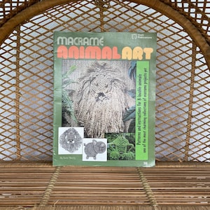 May include: A vintage macrame book titled "Macrame Animal Art" with a green cover. The cover features a macrame lion, pig, and frog. The book is displayed on a woven wicker chair.