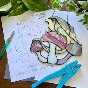 Könnte beinhalten: Ein Drache aus Buntglas, der auf einem Pilz sitzt, mit schillerndem Glas und weißem Hintergrund. Das Bild zeigt auch ein Muster für das Buntglasprojekt, eine Zange und einen Glasschneider.