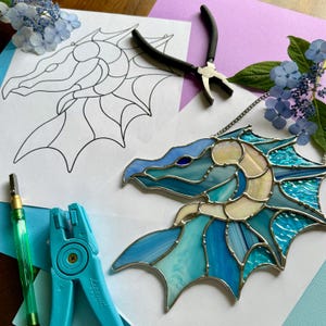 May include: A stained glass dragon head in shades of blue, turquoise, and cream, with a silver metal border. A paper pattern and tools for glasswork are also visible, along with blue flowers.