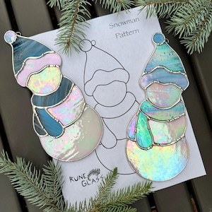 May include: Two stained glass snowman ornaments with iridescent glass, silver edging, and blue, pink, and white accents. A paper pattern with the text "Snowman Pattern" and a snowman outline is behind the ornaments. The ornaments are on a dark wood surface with pine branches.
