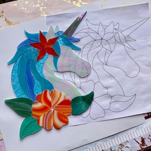 May include: A colorful stained glass unicorn head with a blue mane, a red flower, and an orange and white flower. A line drawing of a unicorn head is next to the stained glass piece.