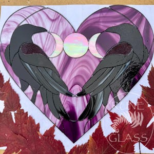 May include: A heart-shaped stained glass panel featuring two black ravens facing each other. Between the birds are three circular moon phases in iridescent glass. The background is composed of swirling shades of purple and white glass.
