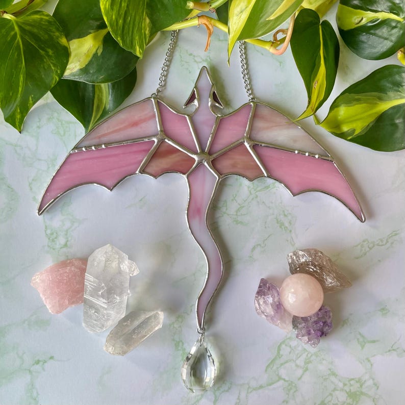 May include: Pink stained glass dragon with a clear crystal teardrop hanging from its tail. The dragon is flying with its wings spread wide. The dragon is surrounded by small pink and clear crystals.