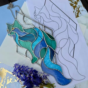 Op de afbeelding: Een glazen kattenhanger in blauwe en groene tinten, met een zilveren ketting. Het kattenontwerp is geplaatst in een geometrische vorm. Een tekening van de kat is zichtbaar op de achtergrond, samen met een boeket paarse bloemen.