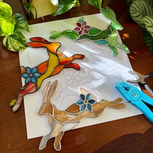 Pode incluir: Quatro figuras de coelho em vitral em várias cores, incluindo laranja, amarelo, verde e castanho, cada uma com um detalhe floral. Um desenho de um coelho é visível por baixo, juntamente com ferramentas.