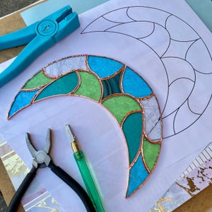 May include: A crescent moon stained glass piece featuring blue, green, and white textured glass segments joined by copper foil. The piece rests on a paper pattern alongside a blue running plier, a black grozing plier, and a glass cutter.