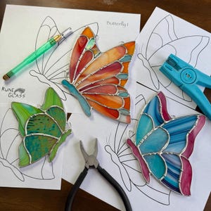 Puede incluir: Tres adornos de mariposa de vidrieras en tonos naranja, verde y azul, con bordes metálicos plateados. Las mariposas están sobre plantillas de patrones de mariposas, con herramientas como alicates y un cortador de vidrio cerca. El texto "Butterfly!" es visible.