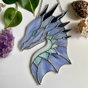 May include: A stained glass dragon head with a blue and teal color scheme. The dragon has a silver chain attached to the top of its head.