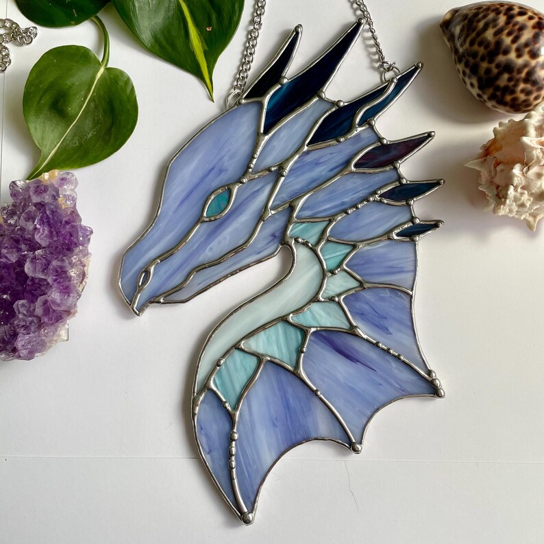 May include: A stained glass dragon head with a blue and teal color scheme. The dragon has a silver chain attached to the top of its head.
