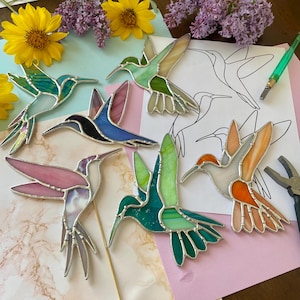 May include: A collection of colorful stained glass hummingbird ornaments in various colors, including green, blue, pink, and orange. Each bird has a silver-colored metal outline. The background includes flowers and a drawing of a hummingbird.