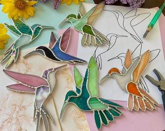 May include: A selection of stained glass hummingbird ornaments in shades of green, blue, pink, and orange. Each bird features a silver metal outline. The background includes flowers and a hummingbird outline drawing.