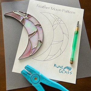 May include: A stained glass moon pattern with a completed stained glass moon, a glass cutter, and a pair of glass nippers. The pattern is labeled "Feather Moon Pattern" and the completed moon is pink and iridescent. The pattern is from Rune Glass.
