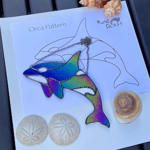 May include: A stained glass orca pendant with a chain, featuring iridescent blues, greens, and purples. The pendant is displayed with a printed "Orca Pattern" and "Rune Glass" design, along with seashells and sand dollars.