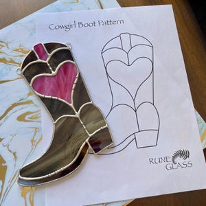 May include: A stained glass cowgirl boot with a pink heart design, set against a printed "Cowgirl Boot Pattern" and a decorative background. The boot features brown and gray glass with a silver metal border. The Rune Glass logo is visible.