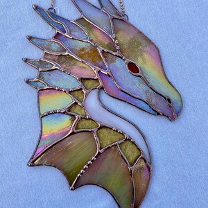 May include: A stained glass dragon head with iridescent colors, including blue, green, yellow, and pink. The dragon's eye is red, and the wings are spread out. The dragon is hanging from a chain.