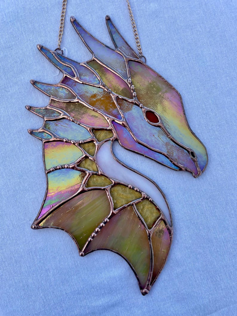 May include: A stained glass dragon head with iridescent colors, including blue, green, yellow, and pink. The dragon's eye is red, and the wings are spread out. The dragon is hanging from a chain.