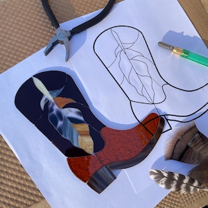 Puede incluir: Una bota de vaquero de vidrieras con un diseño de plumas en tonos azules, marrones y ámbar. La bota se coloca sobre una superficie blanca con un contorno negro de una bota, unos alicates y una herramienta verde.