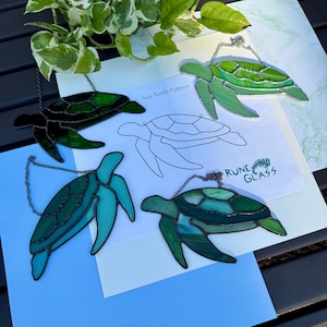 Puede incluir: Adornos de tortugas marinas de vidrieras en tonos verdes y azules, cada uno con una cadena para colgar. También se ve un papel con un patrón de tortuga marina y el texto "Rune Glass".