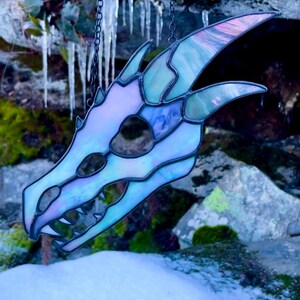 A stained glass dragon skull with iridescent white and grey glass. The skull is hanging from a chain against a background of rocks and icicles. The piece is finished in black patina.