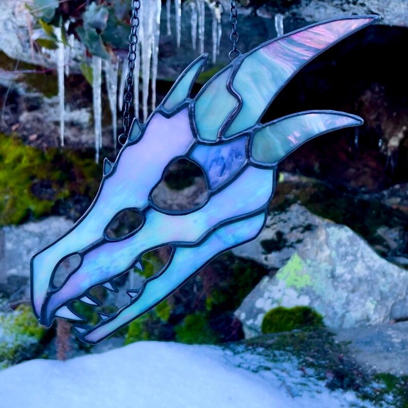 A stained glass dragon skull with iridescent white and grey glass. The skull is hanging from a chain against a background of rocks and icicles. The piece is finished in black patina.
