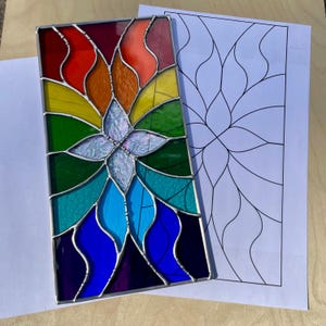 May include: A rectangular stained glass panel featuring a central iridescent white flower shape surrounded by a vibrant rainbow spectrum of red, orange, yellow, green, teal, blue, and purple glass segments held together by dark metal solder lines.