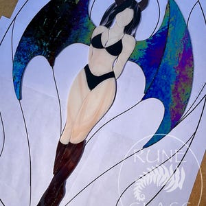 May include: A stained glass art piece depicting a figure with dark hair, horns, and large iridescent blue and purple wings. The figure wears a black bikini and dark brown boots. The piece is placed on a paper pattern with the text RUNE GLASS.