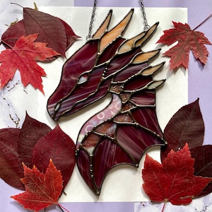 May include: A stained glass dragon head with a burgundy and orange color scheme. The dragon head is hanging from a black chain and is surrounded by red and orange leaves.
