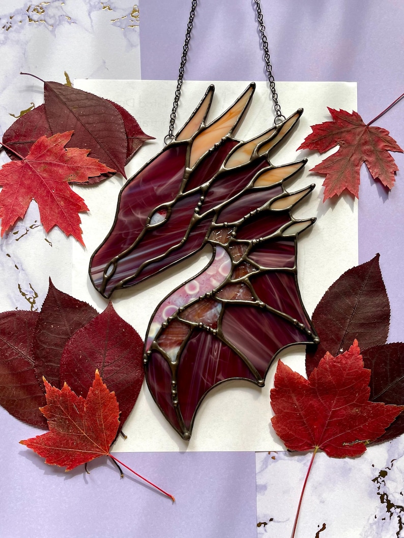 May include: A stained glass dragon head with a burgundy and orange color scheme. The dragon head is hanging from a black chain and is surrounded by red and orange leaves.