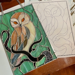 May include: A stained glass owl with orange eyes and brown, tan, and cream-colored feathers, perched on a black branch against a green background. The artwork is framed with a silver metal border and a chain for hanging. A paper pattern is next to the artwork.