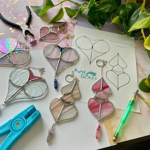 May include: A collection of stained glass heart-shaped ornaments with different patterns and colors, including pink, white, and red. The ornaments are hanging from chains and have crystals attached to the bottom. The image also includes a sheet of paper with the text "Booty and Bust Patterns" and the logo "Rune Glass".