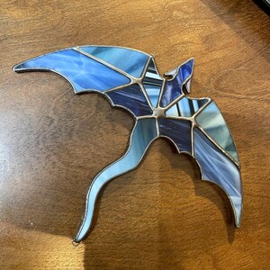 May include: A stained glass dragon with a blue and purple color scheme. The dragon is flying with its wings spread out and its tail curled behind it.