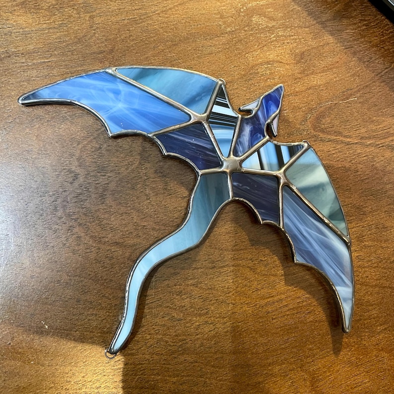 May include: A stained glass dragon with a blue and purple color scheme. The dragon is flying with its wings spread out and its tail curled behind it.
